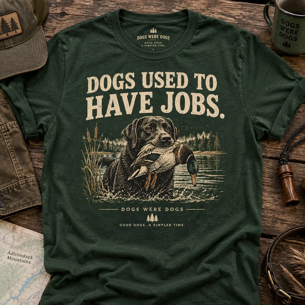 Dogs Used to Have Jobs t-shirt in heather forest green, printed with a retriever carrying a mallard through lake shallows, laid flat on weathered wood beside a waxed cap and an enamel camp mug.