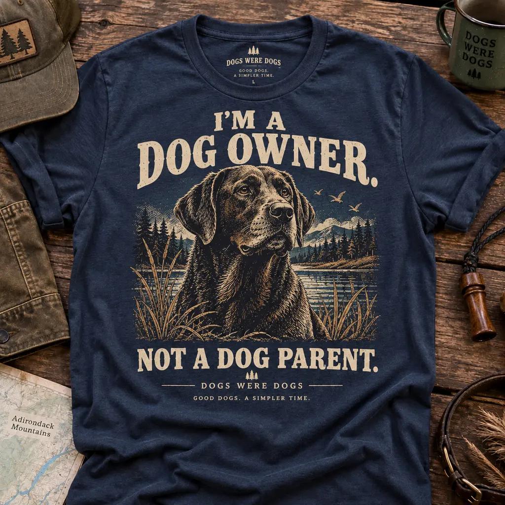 Dog Owner, Not a Dog Parent t-shirt in weathered navy, printed with a seated Labrador portrait against pines and a mountain lake, laid flat on weathered wood.