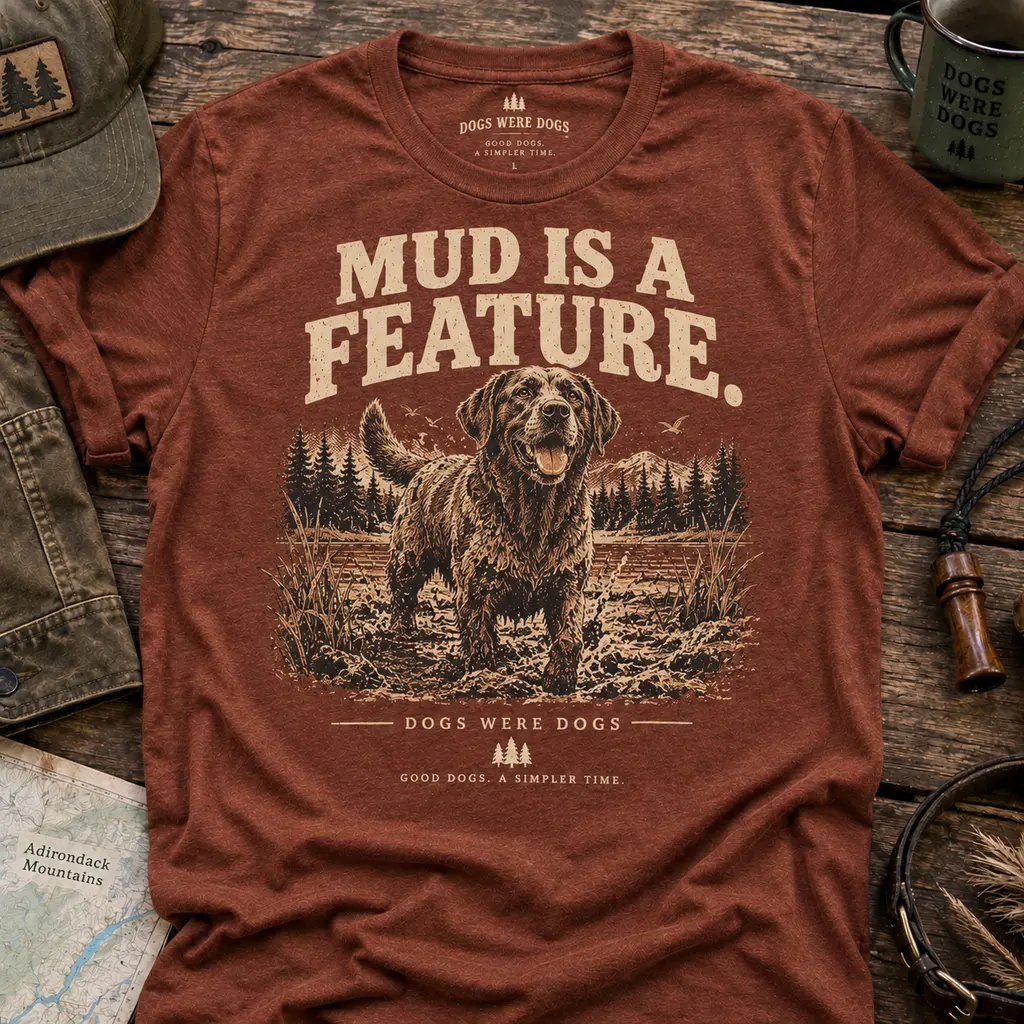 Mud Is a Feature t-shirt in heather rust, printed with a filthy, delighted retriever standing in a churned muddy field, laid flat on weathered wood.