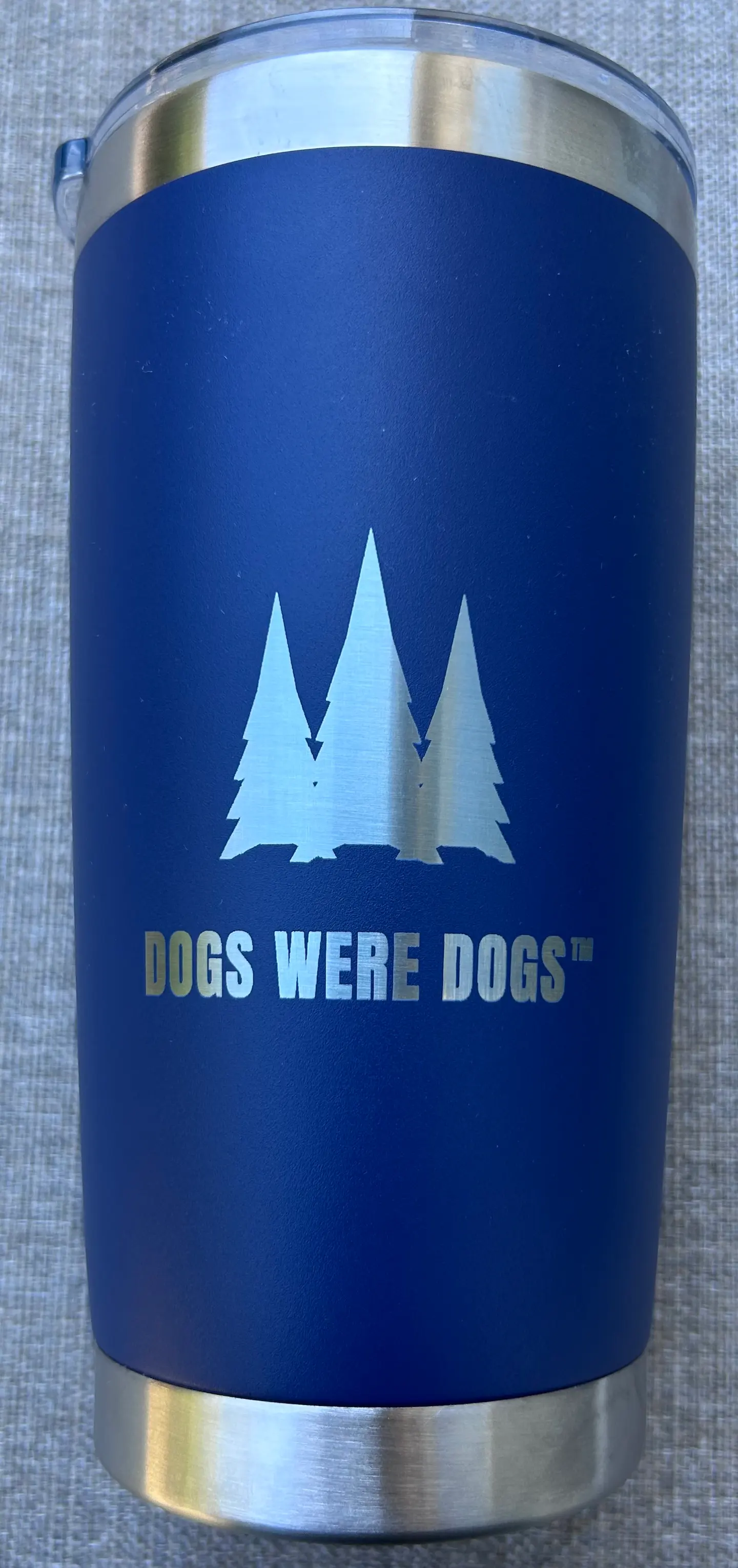 Dog Owner Tumbler, 20 fl oz insulated stainless steel in powder-coated navy, shown front-forward with three laser-engraved pines above the DOGS WERE DOGS wordmark, standing on a woven gray surface.